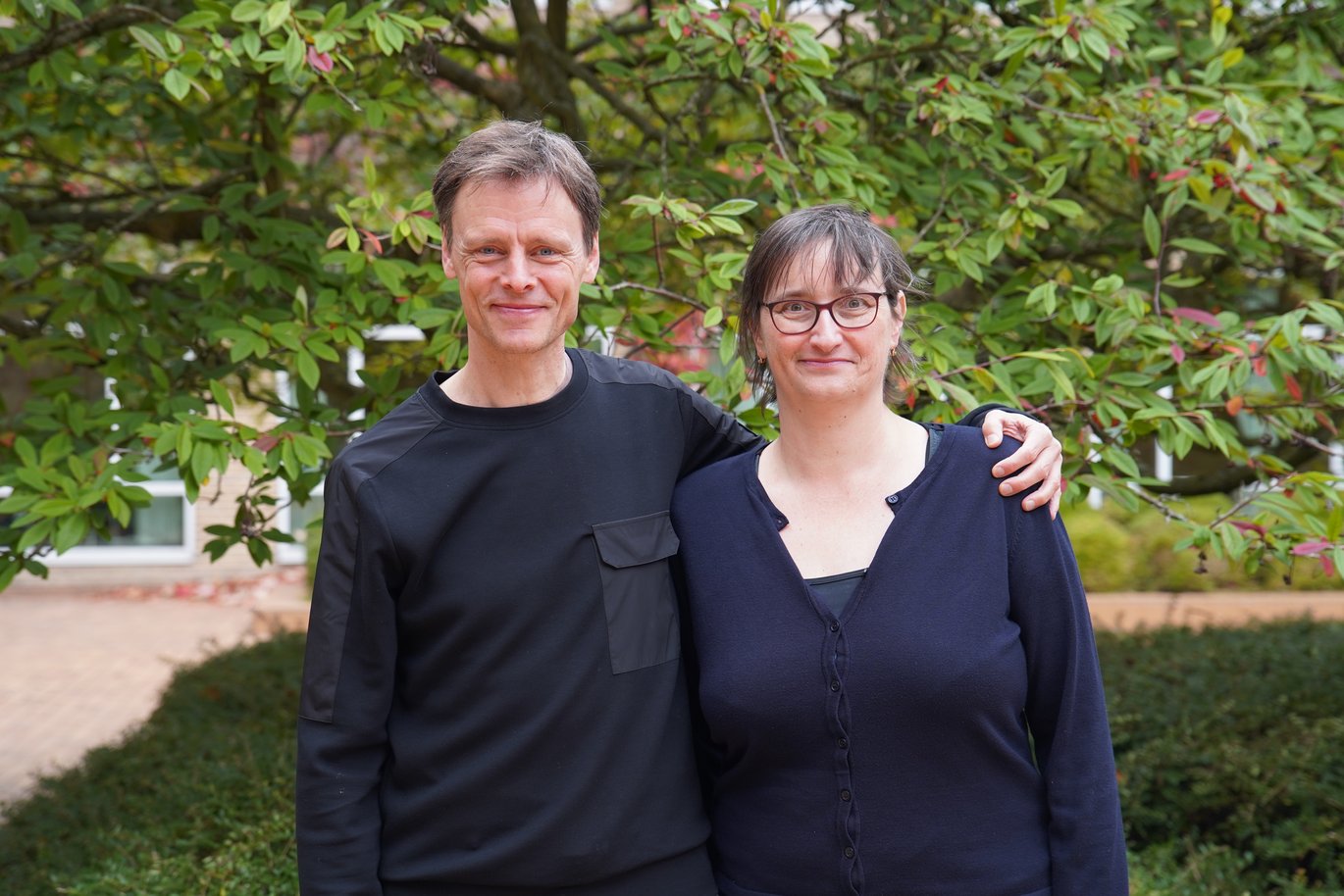 Portrait of researchers Asger Hobolth and Victoria Birkedal standing outdoors in front of green foliage, smiling at the camera. They co-lead a VILLUM Synergy project at Aarhus University.
